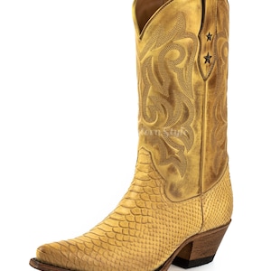 Könnte beinhalten: Ein goldgelber Cowboy-Stiefel mit spitzer Zehenpartie und einem gestapelten Absatz. Der Stiefel hat ein strukturiertes, schlangenartiges Muster am unteren Teil und dekorative Nähte am oberen Schaft. Zwei kleine sternförmige Akzente sind oben sichtbar.