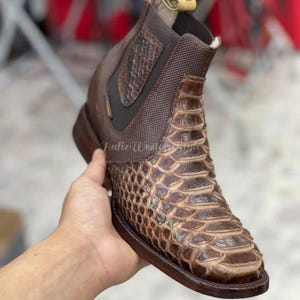 Puede incluir: Botín de cuero marrón con un estampado de piel de serpiente en la pala y un panel texturizado en el lateral. El botín tiene una puntera cuadrada y un tacón apilado. El botín está sujeto por una mano.