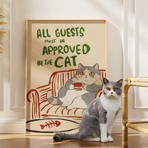 Può includere: Una stampa incorniciata con il testo "ALL GUESTS MUST BE APPROVED BY THE CAT" presenta un gatto dei cartoni animati su un divano a strisce rosse. Un vero gatto con pelo grigio e bianco è seduto davanti alla stampa. Un vaso con piante secche è a sinistra.