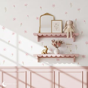 May include: Two pink scalloped wall shelves mounted on a wall with pink floral decals. The top shelf holds a gold-framed mirror, a framed botanical print, and a plush rabbit. The bottom shelf holds a gold rabbit figurine, a vase of dried flowers, and a jewellery stand.