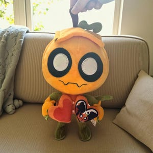 May include: Orange plush toy with large black and white eyes, a stitched mouth, and a green stem. The toy holds two heart-shaped objects, one red and one with a black and white design. The toy is on a beige couch.