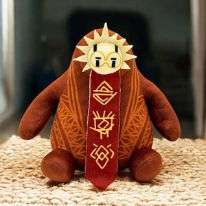 May include: A brown and orange plush toy with a sun-shaped head, glasses, and a red scarf with gold symbols. The toy has a rounded body, small arms, and feet, and is sitting on a textured surface.