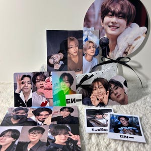 May include: A collection of K-pop idol merchandise featuring multiple rectangular photo cards, a circular fan with a portrait, and two square polaroid-style prints. The items display various portraits of a young person with dark hair, some with text like EN-.