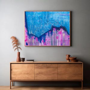 May include: A rectangular framed painting featuring a vibrant abstract composition with a dominant blue upper section and a bright pink lower section, separated by dark jagged lines. The artwork is displayed on a wall above a wooden sideboard.