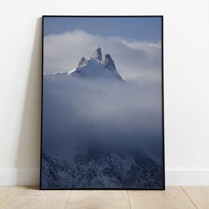Op de afbeelding: Een ingelijste print met een besneeuwde bergtop die uit wolken oprijst. De afbeelding is zwart omlijst en geplaatst tegen een witte muur en een houten vloer.