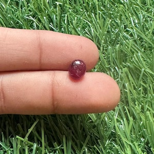 Può includere: Una piccola gemma rosso scuro, lucidata e di forma ovale. La gemma è tenuta tra due dita su uno sfondo di erba artificiale verde. La gemma è di un colore ricco e traslucido.