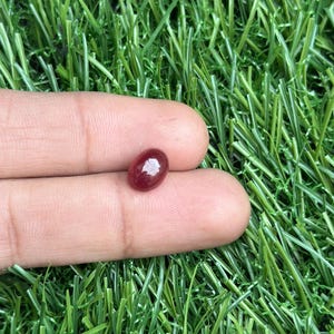 Może przedstawiać: Mały, owalny, wypolerowany kamień szlachetny w głębokim, czerwonobrązowym odcieniu. Klejnot trzymany jest między dwoma palcami na tle zielonej sztucznej trawy. Powierzchnia kamienia odbija światło, podkreślając jego gładką fakturę.