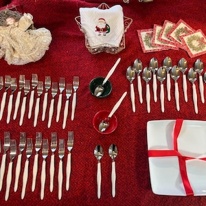 May include: A Christmas-themed table setting with cutlery, plates, and decorations. The arrangement includes forks, spoons, and a square plate with a red ribbon. A napkin with a Santa Claus design is in a star-shaped holder.