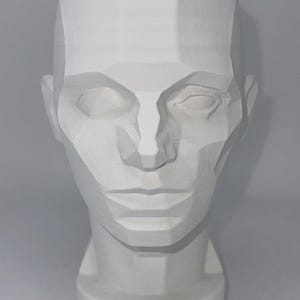 May include: A white, matte-finished sculptural head bust featuring a stylized, geometric, and faceted design. The facial features, including the eyes, nose, and lips, are defined by sharp, angular planes and flat surfaces, creating a structured appearance.