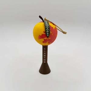 May include: A decorative Christmas ornament shaped like a peach. The ornament is yellow with red accents and a green leaf. The base is brown with the word "CLANTON" printed on it. A gold string is attached for hanging.