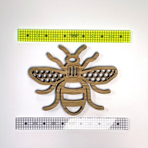 May include: A wooden cutout of a bee with a honeycomb pattern on its wings. The bee is brown and has a black outline. The bee is approximately 2 inches wide.