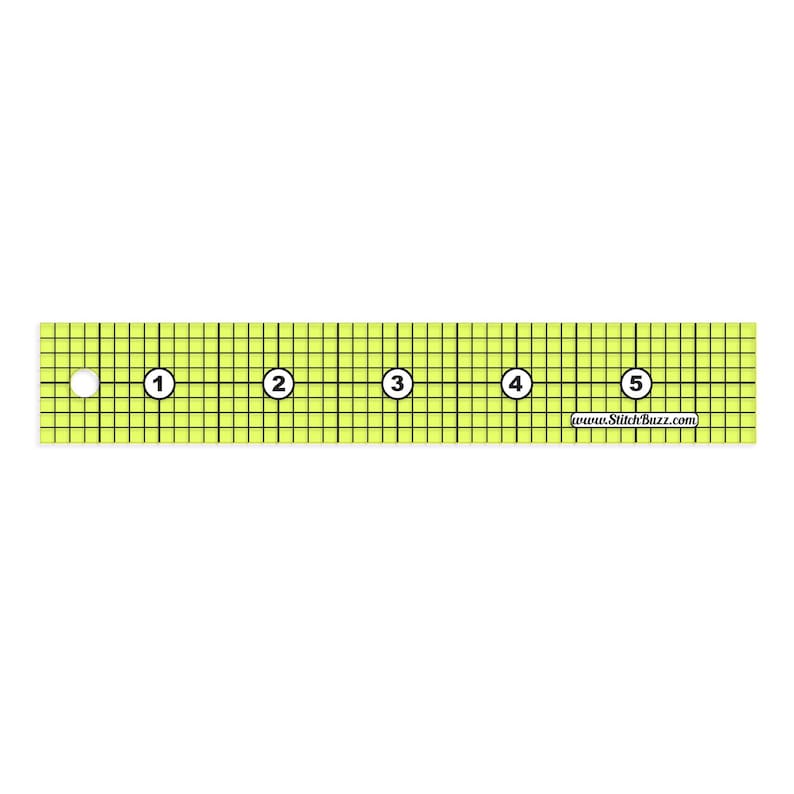 May include: A yellow ruler with black markings and numbers 1 through 5. The ruler is divided into small squares. The text "www.StitchBuzz.com" is printed on the ruler.