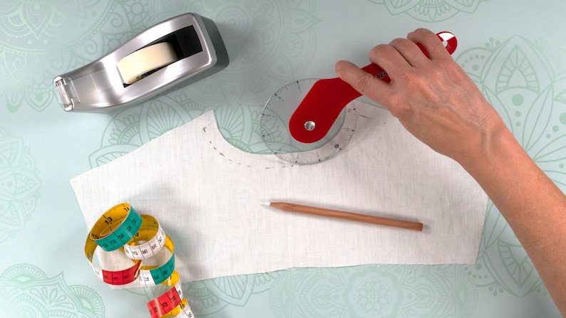 Puede incluir: Una escena de costura con un trozo de tela blanca marcado con un cortador rotatorio rojo. Tambi&eacute;n hay un dispensador de cinta adhesiva, un l&aacute;piz y una cinta m&eacute;trica, lo que sugiere la confecci&oacute;n de prendas.