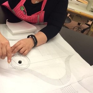 Puede incluir: Una persona utiliza un transportador de pl&aacute;stico transparente para medir un patr&oacute;n en papel blanco. El patr&oacute;n tiene l&iacute;neas curvas. Una m&aacute;quina de coser est&aacute; en el fondo. La persona lleva una camisa negra de manga larga y un delantal rosa.