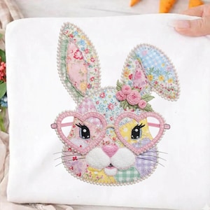 May include: A white fabric square featuring a patchwork bunny face with heart-shaped glasses. The bunny has floral patterned ears and a pink nose. The glasses are pink and the bunny is decorated with small pink roses and a pearl border.