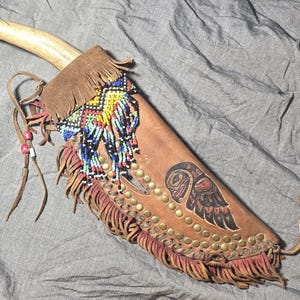 Könnte beinhalten: Ein handgefertigtes Messer mit hellem Griff und Lederscheide. Die Scheide ist mit farbenfrohen Perlenarbeiten, Fransen und einem gemalten Eulenmotiv verziert. Die Scheide ist braun mit Messingnieten.