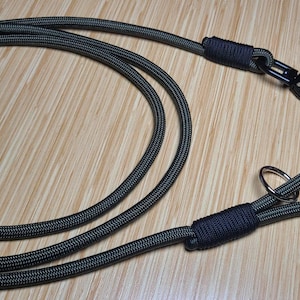 May include: A dark olive green rope leash featuring a black metal swivel snap hook at one end and a looped handle at the other. The connection points are secured with tightly wrapped black cord, and a silver metal ring is attached near the handle.