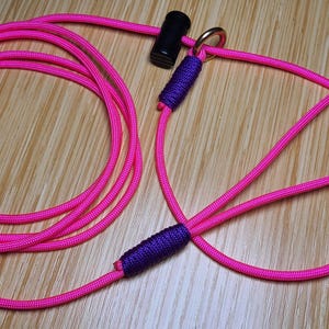 May include: A bright neon pink paracord slip lead for pets, featuring a black cylindrical stopper, a metallic ring, and two sections of purple cord wrapping that secure the loops. The long, flexible rope is coiled into a circular shape.