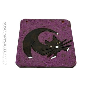 Puede incluir: Una placa cuadrada de color morado con una superficie texturizada que presenta la silueta de un gato negro arqueado sobre una forma de media luna. El gato tiene patas con puntas blancas, nariz blanca y ojos verdes. El texto SELECTEDBYSANNEDSGN está impreso verticalmente.
