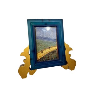 Peut inclure: Petite œuvre d'art encadrée représentant une scène de pont, dans un cadre bleu sarcelle. Le cadre est soutenu par un support décoratif doré. L'œuvre représente un pont sur l'eau avec des personnages.