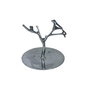 Op de afbeelding: Een zilverkleurige metalen decoratieve standaard met een boomachtig ontwerp en een vogel die op een tak zit. De standaard heeft een cirkelvormige basis, geschikt voor het tentoonstellen van kleine items.