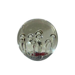May include: A clear glass paperweight sphere featuring internal suspended air bubbles arranged in a vertical pattern. The base of the sphere contains a dark red accent and a textured silver metallic layer that reflects light through the glass.