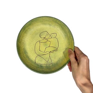 May include: A round, green decorative plate with a hand-drawn illustration of a person holding a child. The plate has a textured surface and a dark green rim. The illustration is in black.
