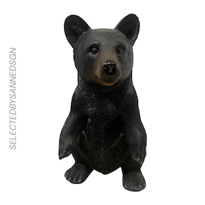 Peut inclure: Une figurine d'ours noir debout sur ses pattes arrière avec une finition en fourrure texturée, des yeux sombres et un museau teinté de brun. Le texte SELECTEDBYSANNEDSGN est imprimé verticalement en gris clair sur le côté gauche.