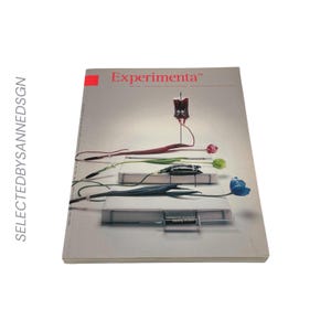 May include: A softcover magazine titled Experimenta 54 with a light grey cover. The graphic design features three white rectangular blocks, each connected to a medical IV bag, a syringe, or a tube, with colourful tulips extending from the objects.