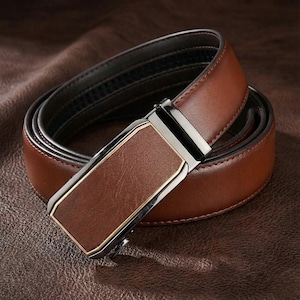 Op de afbeelding: Een bruine leren riem met een zilveren gesp en een goudkleurige rand. De riem is opgerold, wat de gladde textuur en het ontwerp laat zien. De gesp is rechthoekig.