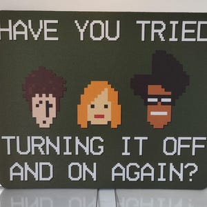 May include: A rectangular dark olive green sign featuring white pixelated text that reads HAVE YOU TRIED TURNING IT OFF AND ON AGAIN? Below the first line of text are three pixelated character heads with brown, orange, and dark brown hair.