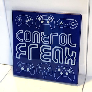 May include: A square blue sign featuring white line art of six different video game controllers arranged around the text "control freak" written in a bold, stylised, geometric white font centred on the surface of the glossy blue plaque.