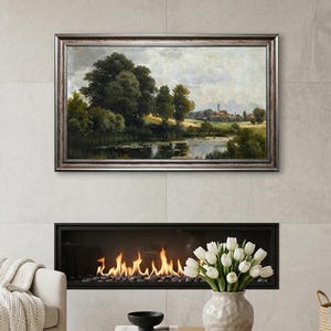 May include: A framed landscape painting depicting a serene waterside scene with trees and a distant village. The painting hangs above a fireplace with a modern design. A beige armchair, a round wooden coffee table, and a vase of white tulips complete the room.