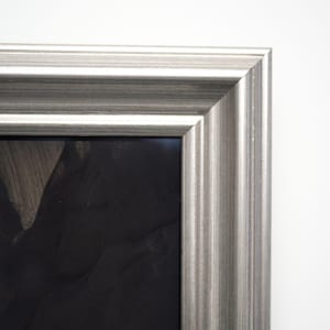 Puede incluir: Marco de fotos plateado con textura acanalada. El marco tiene un diseño simple y elegante y un color neutro que complementa varios estilos de obras de arte. La esquina del marco es visible sobre un fondo blanco.