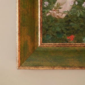 Op de afbeelding: Close-up van een ingelijst schilderij met een groene en gouden houten lijst. Het kunstwerk toont een hand die een tak met roze bloemen en groene bladeren vasthoudt. De lijst heeft een verweerde, antieke uitstraling.