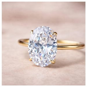 May include: A gold ring with a large, oval-shaped diamond. The diamond is clear and cut to reflect light. The ring has a plain, polished gold band. The diamond is held by four prongs. The background is a soft, neutral colour.