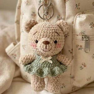 Könnte beinhalten: Ein gehäkelter Teddybär-Schlüsselanhänger in Beige mit einem grünen Kleid und einer weißen Schleife. Der Bär hat eine braune Nase, schwarze Augen und rosa Wangen. Der Schlüsselanhänger ist an einem silbernen Verschluss befestigt.