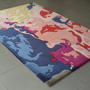 May include: Rectangular area rug with an abstract design in shades of blue, pink, red, and beige. The rug's pattern features irregular shapes and forms, creating a modern and artistic aesthetic. The rug is placed on a tiled floor.