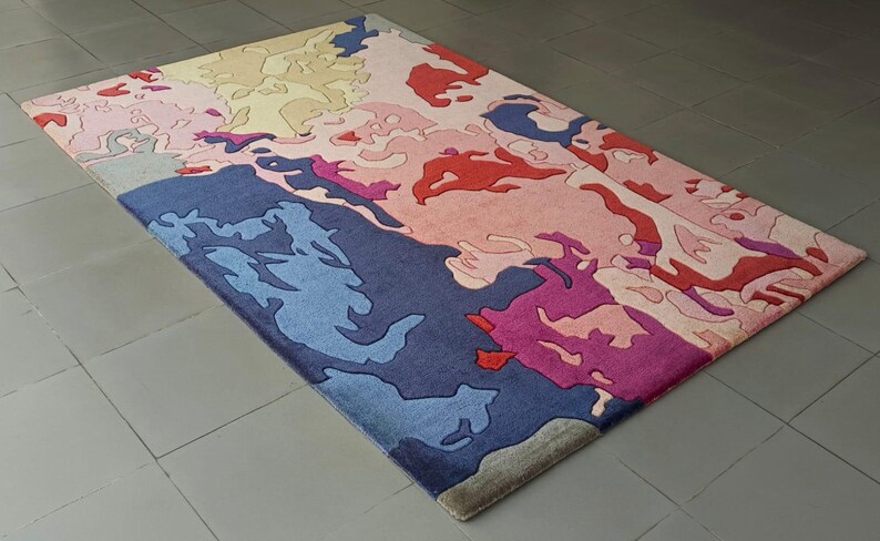 May include: Rectangular area rug with an abstract design in shades of blue, pink, red, and beige. The rug's pattern features irregular shapes and forms, creating a modern and artistic aesthetic. The rug is placed on a tiled floor.