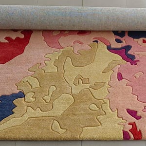 May include: A rolled-up area rug with an abstract design. The rug features a mix of colors, including pink, red, yellow, blue, and gray. The design has a map-like appearance with irregular shapes and patterns. The rug is made of a soft, plush material.