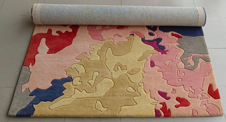 May include: A rolled-up area rug with an abstract design. The rug features a mix of colors, including pink, red, yellow, blue, and gray. The design has a map-like appearance with irregular shapes and patterns. The rug is made of a soft, plush material.