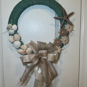 May include: A decorative wreath with a teal green burlap base. Adorned with seashells, faux flowers, a starfish, and a large bow made of cream and tan ribbons. The wreath is designed for coastal or beach-themed decor.