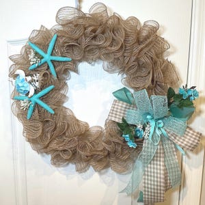 May include: A decorative wreath made of brown mesh, adorned with turquoise starfish and a bow. The bow features a gingham pattern and is accented with turquoise ribbons and small blue flowers. This wreath is perfect for coastal or beach-themed decor.