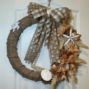 May include: A circular wreath wrapped in brown burlap, featuring a large bow with tan and white gingham and polka dot ribbons. It is decorated with a white starfish, a white sand dollar, a small white turtle figure, and tan artificial flowers.