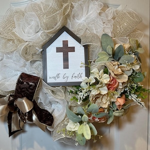 May include: A decorative wreath featuring a sign with a cross and the words "walk by faith." The wreath is made of beige and white mesh, with a brown ribbon and floral accents in peach, green, and white.