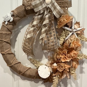 May include: A circular wreath wrapped in tan burlap with a twine spiral. It features a beige and white gingham bow, a white turtle figure, a white starfish, a white sand dollar, various tan and brown artificial flowers, and small seashells.