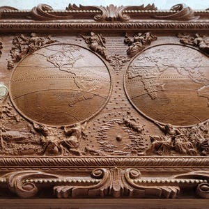 Op de afbeelding: Een gedetailleerde houten kist met een gebeeldhouwd hemels thema. Het heeft een zon, maan, globes en navigatie-instrumenten. De kist is bruin met sierlijke krullen aan de randen en een messing kompas en telescoop.