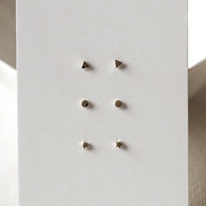 May include: A set of six gold stud earrings on a white card. The earrings are in the shapes of a triangle, circle, and star.