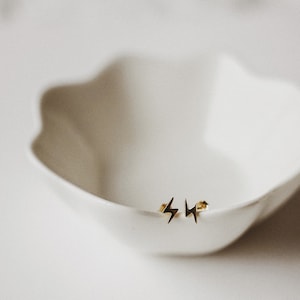 Puede incluir: Un par de pendientes de bot&oacute;n dorados con forma de rayo. Los pendientes son peque&ntilde;os y delicados, con un dise&ntilde;o simple y minimalista. Se colocan en el borde de un cuenco blanco con borde festoneado. El fondo es de un color suave y neutro.