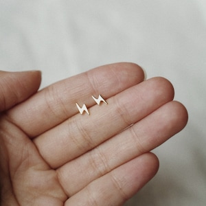 Puede incluir: Un par de peque&ntilde;os pendientes de tuerca dorados con forma de rayo. Los pendientes se muestran sobre un fondo claro, mostrando su dise&ntilde;o minimalista. Los pendientes se sostienen en una mano.